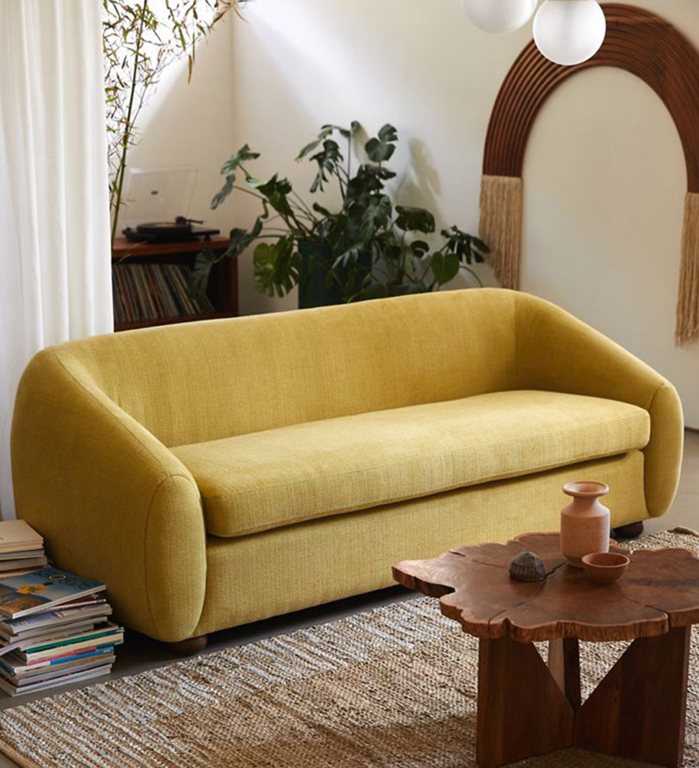 A modern curved yellow fabric sofa in a styled living room setting with a wooden coffee table.